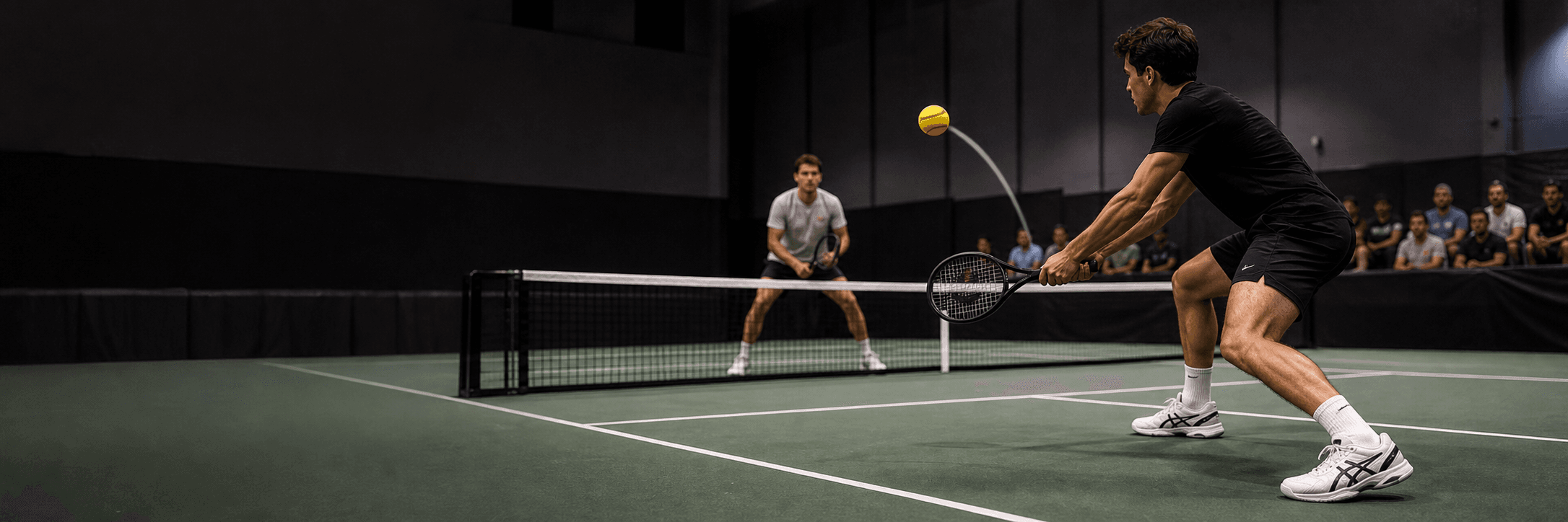Two TYPTI players rally across a low net on a dark indoor court as a yellow foam ball flies mid-flight off a carbon-fiber racket.